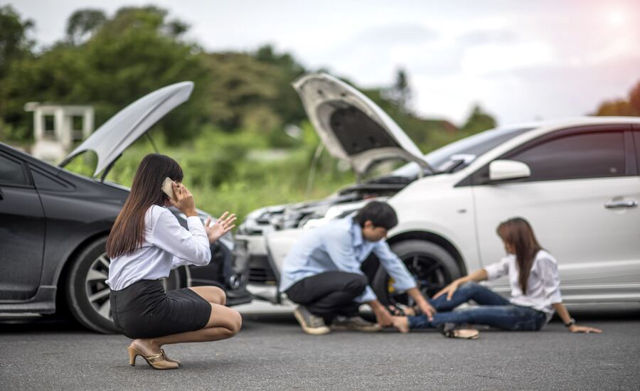 An injured woman sitting on the ground after a car accident, with a man helping her and another woman calling an ambulance.